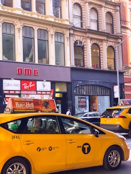 A busy urban street scene featuring a yellow NYC taxi with an advertisement on top. The street is lined with historic buildings housing stores, including Calvin Klein and Ray-Ban, with sale signs displayed in the windows. Multiple pedestrians and cars are visible, contributing to the bustling atmosphere.