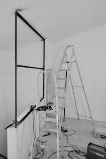 A black and white photograph depicting a construction or renovation scene. Two ladders, one taller and another shorter, are placed within an unfinished room that features a partially built wall frame. Various tools, including a drill and a tool box, are scattered on the floor, along with some cables. The environment is minimalistic and appears to be an interior space under construction.