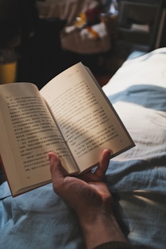 A hand is holding an open book with text in a script that appears to be Chinese. The book is being read in what seems to be a cozy setting, possibly on a bed or couch, with a blanket partially covering the person holding the book. The lighting is soft and warm, suggesting a relaxing atmosphere.