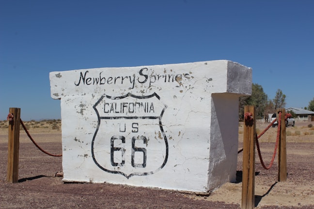 A weathered cement sign with the Route 66 shield and writings 'Newberry Springs' and 'California US 66' is positioned in a desert landscape. It is supported by wooden posts connected with red ropes, with clear blue skies and dry vegetation in the background.