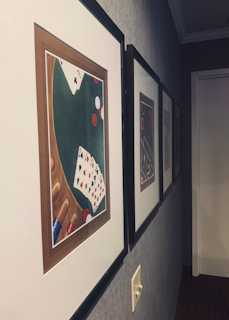 A hallway features framed paintings on the wall, depicting a poker game with playing cards and poker chips on a green table. The lighting switch is visible on the wall underneath the paintings.