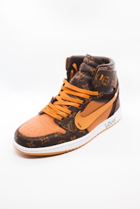 A stylish high-top sneaker featuring a combination of brown and orange tones with a luxurious pattern. The shoe has orange laces and a prominent swoosh logo on its side. The material appears textured, giving off an expensive and designer look. The sneaker is positioned against a plain, light background, highlighting its design.