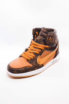 A stylish high-top sneaker featuring a combination of brown and orange tones with a luxurious pattern. The shoe has orange laces and a prominent swoosh logo on its side. The material appears textured, giving off an expensive and designer look. The sneaker is positioned against a plain, light background, highlighting its design.