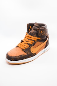 A stylish high-top sneaker featuring a combination of brown and orange tones with a luxurious pattern. The shoe has orange laces and a prominent swoosh logo on its side. The material appears textured, giving off an expensive and designer look. The sneaker is positioned against a plain, light background, highlighting its design.