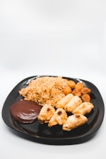 A black plate containing an assortment of foods, including a pile of fried rice, several spring rolls or pastries, round fried snacks, and a portion of brown sauce or dip.