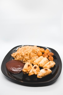 A black plate containing an assortment of foods, including a pile of fried rice, several spring rolls or pastries, round fried snacks, and a portion of brown sauce or dip.