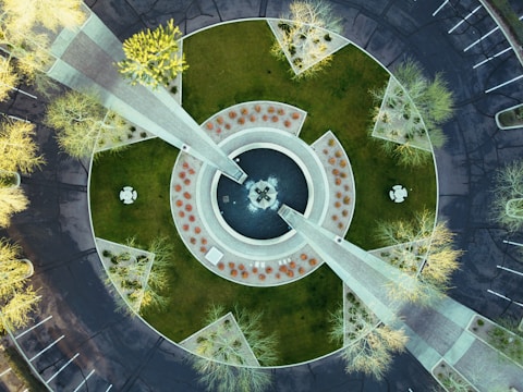 An aerial view of a circular plaza with geometric landscaping, featuring pathways, grass, and trees. The design includes concentric circles with a central fountain area surrounded by neatly arranged shrubbery and paving. Sparse trees cast long shadows across the paved areas, and parking lots with white lines are visible on the outskirts.