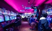 A vibrant arcade with rows of gaming machines adorned with colorful screens. Several people are immersed in playing games, seated on stools in a dimly lit room accented by an intense pink ceiling light. The atmosphere is lively and engaging.
