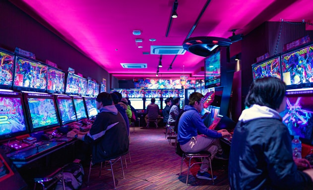 A vibrant arcade with rows of gaming machines adorned with colorful screens. Several people are immersed in playing games, seated on stools in a dimly lit room accented by an intense pink ceiling light. The atmosphere is lively and engaging.