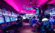 A vibrant arcade with rows of gaming machines adorned with colorful screens. Several people are immersed in playing games, seated on stools in a dimly lit room accented by an intense pink ceiling light. The atmosphere is lively and engaging.