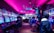 A vibrant arcade with rows of gaming machines adorned with colorful screens. Several people are immersed in playing games, seated on stools in a dimly lit room accented by an intense pink ceiling light. The atmosphere is lively and engaging.
