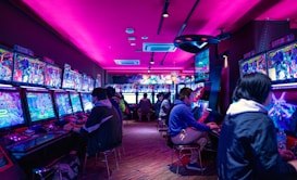 A vibrant arcade with rows of gaming machines adorned with colorful screens. Several people are immersed in playing games, seated on stools in a dimly lit room accented by an intense pink ceiling light. The atmosphere is lively and engaging.
