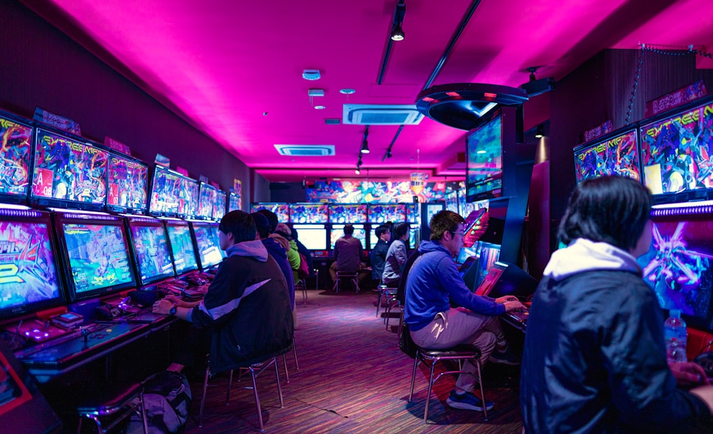 A vibrant arcade with rows of gaming machines adorned with colorful screens. Several people are immersed in playing games, seated on stools in a dimly lit room accented by an intense pink ceiling light. The atmosphere is lively and engaging.