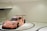 A sports car with a vibrant pink livery featuring various sponsor logos is displayed in a minimalistic, modern showroom with white walls and a glossy floor. The car has a prominent spoiler and racing decals, giving it a dynamic appearance.