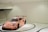 A sports car with a vibrant pink livery featuring various sponsor logos is displayed in a minimalistic, modern showroom with white walls and a glossy floor. The car has a prominent spoiler and racing decals, giving it a dynamic appearance.