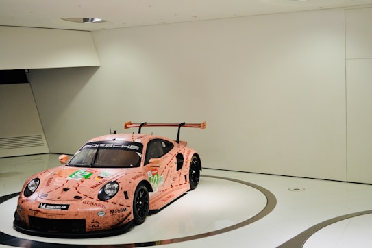 A sports car with a vibrant pink livery featuring various sponsor logos is displayed in a minimalistic, modern showroom with white walls and a glossy floor. The car has a prominent spoiler and racing decals, giving it a dynamic appearance.