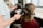 A person is getting their hair styled with a blow dryer and a round brush. The individual is seated, wearing a maroon top, while another person holds the styling tools, focusing on creating curls or volume. The environment resembles a hair salon with professional equipment.