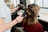 A person is getting their hair styled with a blow dryer and a round brush. The individual is seated, wearing a maroon top, while another person holds the styling tools, focusing on creating curls or volume. The environment resembles a hair salon with professional equipment.
