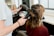 A person is getting their hair styled with a blow dryer and a round brush. The individual is seated, wearing a maroon top, while another person holds the styling tools, focusing on creating curls or volume. The environment resembles a hair salon with professional equipment.