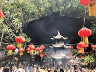 A bustling scene at a traditional outdoor event, featuring numerous red lanterns hanging from strings amidst green foliage. A small pagoda-like structure is at the center, surrounded by a crowd of people, many of whom are holding incense sticks. The atmosphere appears festive and vibrant, with smoke rising into the air from the incense.