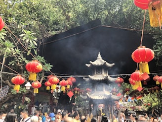 A bustling scene at a traditional outdoor event, featuring numerous red lanterns hanging from strings amidst green foliage. A small pagoda-like structure is at the center, surrounded by a crowd of people, many of whom are holding incense sticks. The atmosphere appears festive and vibrant, with smoke rising into the air from the incense.
