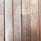 Wooden planks aligned vertically, featuring a natural wood grain and small evenly spaced screws securing them in place. The texture and color of the wood suggest aging, with slight wear and discoloration visible on the surface.