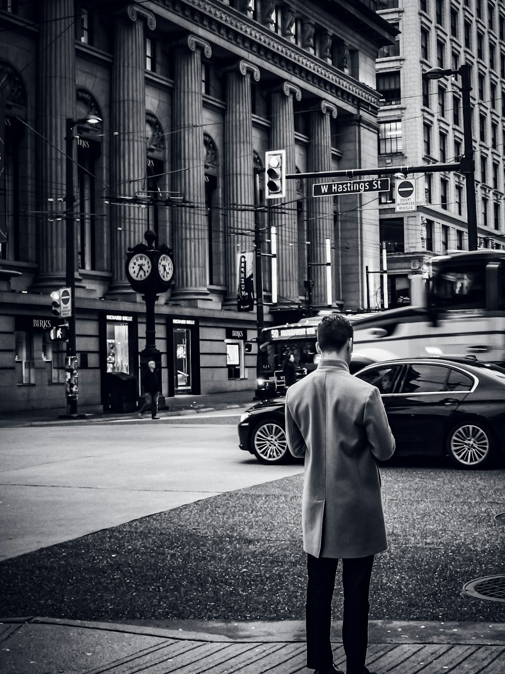 grayscale photo of woman in coat standing on sidewalk near cars and buildings