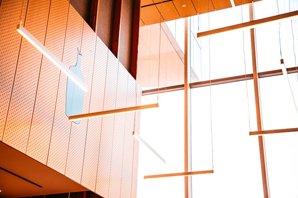 A modern interior with large windows and wooden paneling featuring suspended linear lights. A subtle light blue bottle silhouette is present on a perforated white panel wall.