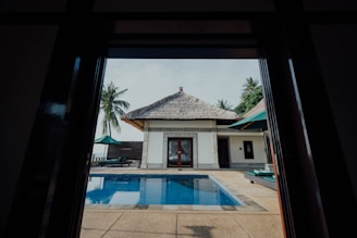 A view of a tropical outdoor area framed by a doorway, featuring a rectangular swimming pool with clear blue water. A single-story white building with a thatched roof is situated opposite the pool. The background includes palm trees and a clear sky with few clouds. There are deck chairs and umbrellas placed around the pool area.