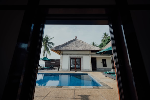 A view of a tropical outdoor area framed by a doorway, featuring a rectangular swimming pool with clear blue water. A single-story white building with a thatched roof is situated opposite the pool. The background includes palm trees and a clear sky with few clouds. There are deck chairs and umbrellas placed around the pool area.