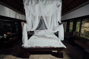 A beautifully arranged bed with white linens and a canopy of sheer white fabric draping elegantly from above. The room is spacious with dark wooden accents, large windows showing a view of greenery, and a touch of natural light filtering in. On the bed are small yellow flowers, adding a touch of color to the clean aesthetic. In the background, there is a fruit basket and other decorative items on a table.