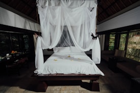 A beautifully arranged bed with white linens and a canopy of sheer white fabric draping elegantly from above. The room is spacious with dark wooden accents, large windows showing a view of greenery, and a touch of natural light filtering in. On the bed are small yellow flowers, adding a touch of color to the clean aesthetic. In the background, there is a fruit basket and other decorative items on a table.