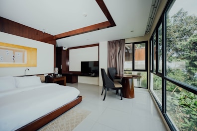 A modern hotel room features a large bed with white linens, a dark wooden headboard, and a bedside table with a lamp. A flat-screen TV is mounted on a cabinet next to a small seating area with two dark chairs and a round wooden table. Large windows provide a view of greenery outside, allowing natural light to flood the room.