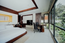 A modern hotel room features a large bed with white linens, a dark wooden headboard, and a bedside table with a lamp. A flat-screen TV is mounted on a cabinet next to a small seating area with two dark chairs and a round wooden table. Large windows provide a view of greenery outside, allowing natural light to flood the room.