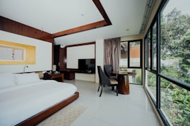 A modern hotel room features a large bed with white linens, a dark wooden headboard, and a bedside table with a lamp. A flat-screen TV is mounted on a cabinet next to a small seating area with two dark chairs and a round wooden table. Large windows provide a view of greenery outside, allowing natural light to flood the room.