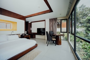 A modern hotel room features a large bed with white linens, a dark wooden headboard, and a bedside table with a lamp. A flat-screen TV is mounted on a cabinet next to a small seating area with two dark chairs and a round wooden table. Large windows provide a view of greenery outside, allowing natural light to flood the room.