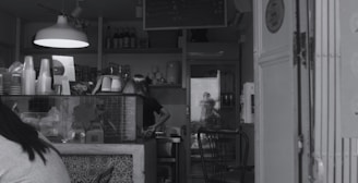 A small cafe with a counter displaying various items, including disposable cups and a menu. A person is working behind the counter, and there is a reflection of another person taking a photo in the background. Shelves with bottles and a hanging light fixture are visible.