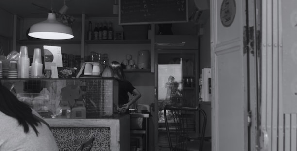 A small cafe with a counter displaying various items, including disposable cups and a menu. A person is working behind the counter, and there is a reflection of another person taking a photo in the background. Shelves with bottles and a hanging light fixture are visible.