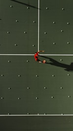 Aerial view of a tennis court with a person in a red shirt standing in the center, surrounded by scattered tennis balls aligned in a grid pattern. The court surface is a deep green with white boundary lines. The person's shadow is clearly visible due to the overhead sunlight.