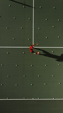 Aerial view of a tennis court with a person in a red shirt standing in the center, surrounded by scattered tennis balls aligned in a grid pattern. The court surface is a deep green with white boundary lines. The person's shadow is clearly visible due to the overhead sunlight.