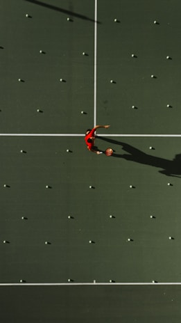 Aerial view of a tennis court with a person in a red shirt standing in the center, surrounded by scattered tennis balls aligned in a grid pattern. The court surface is a deep green with white boundary lines. The person's shadow is clearly visible due to the overhead sunlight.