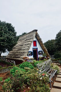 A charming, thatched-roof A-frame house surrounded by a lush garden with colorful flowers and a rustic wooden bench. The house features white walls with blue and red accents, and is situated in a picturesque, green landscape.