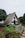 A charming, thatched-roof A-frame house surrounded by a lush garden with colorful flowers and a rustic wooden bench. The house features white walls with blue and red accents, and is situated in a picturesque, green landscape.