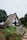 A charming, thatched-roof A-frame house surrounded by a lush garden with colorful flowers and a rustic wooden bench. The house features white walls with blue and red accents, and is situated in a picturesque, green landscape.