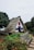 A charming, thatched-roof A-frame house surrounded by a lush garden with colorful flowers and a rustic wooden bench. The house features white walls with blue and red accents, and is situated in a picturesque, green landscape.