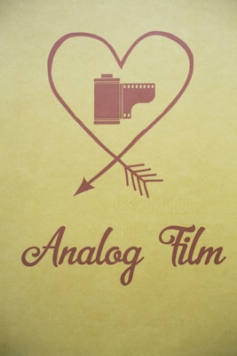 A vintage-inspired design featuring an illustration of a film roll inside a heart shape with an arrow through it. The phrase 'Analog Film' is written in elegant, cursive font below the image. The background is in a muted yellow tone.
