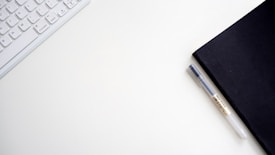 A minimalist workspace setup featuring a white keyboard, a black notebook, and a pen on a plain white surface.