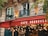 A quaint cafe with a red awning that reads 'Café Beaujolais.' The exterior of the building features ornate architectural details, including arches and decorative stonework. Warm, glowing lights are strung across the front of the cafe, creating a cozy and inviting atmosphere. Greenery is visible, with leaves partially framing the photo.