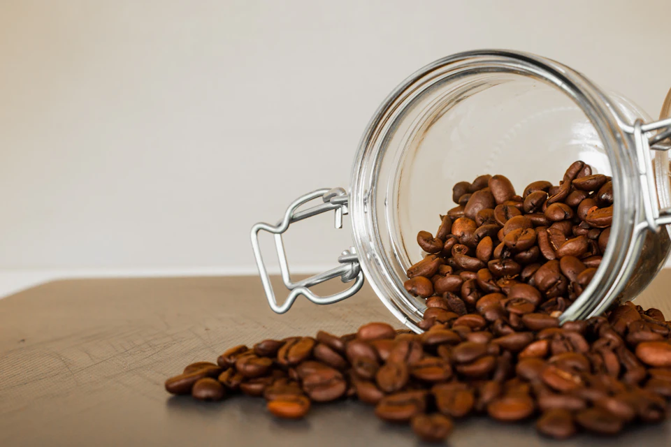 The image shows a glass jar filled with coffee beans spilling out onto a wooden surface, creating an interesting contrast between the transparent jar and the brown coffee beans scattered around it. The jar is placed on its side, allowing the beans to spill over the edge into the surrounding area.
