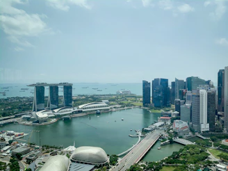 Aerial view of a modern urban landscape with a prominent hotel featuring three towers connected by an elevated platform. Surrounding the hotel are sleek skyscrapers and a body of water housing several small boats. The scene includes lush greenery and a large ferris wheel in the distance.